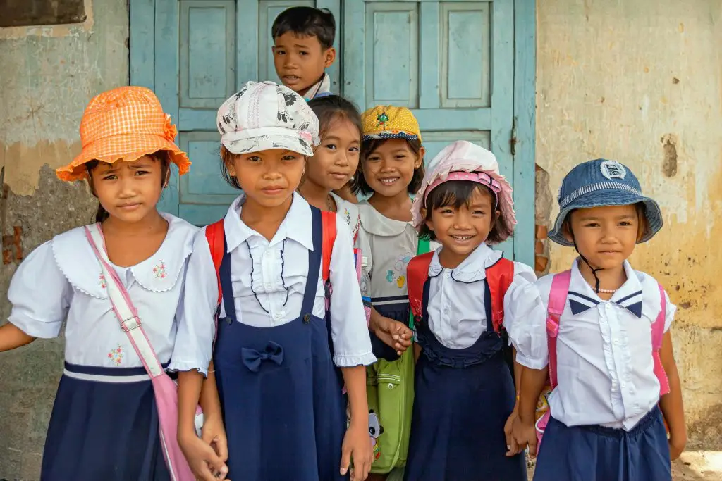 Group of Vietnamese school children smiling in traditional uniforms outside a rustic building.