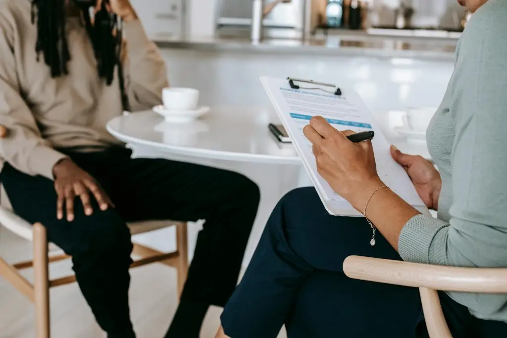 Crop unrecognizable female psychotherapist or interviewer asking questions and taking notes in clipboard during interview with black male